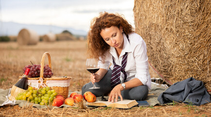 Type representation of freelancer. Girl in blouse and business suit, sitting next to haystack in field, reading book, enjoying day off in country. Woman with glass of wine studies read literature