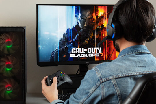 a male gamer at the right side of the picture holding a xbox controller watching a pc monitor with call of duty black ops 7