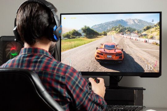 a male gamer at the left side of the picture holding a xbox controller playing forza horizon video game on a computer