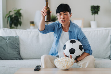 Asian woman holding soccer ball and empty beer bottle while watching tv
