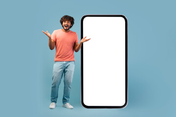 Cool eastern man displays a surprised expression while standing next to a large smartphone with a white screen. He gestures enthusiastically, showcasing a fantastic online opportunity.