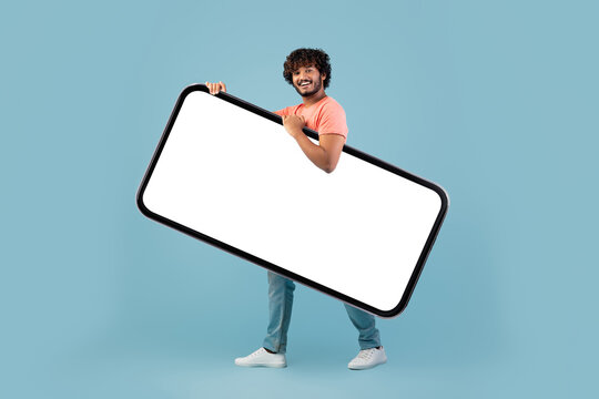 A cheerful Indian man with curly hair is walking while holding a large smartphone with a blank screen. He is in a blue studio setting, showcasing the gadget.