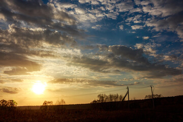 Vibrant sunset sky bursting with golden light and dramatic clouds over a tranquil rural field. Silhouetted trees and power lines mark a peaceful spring evening