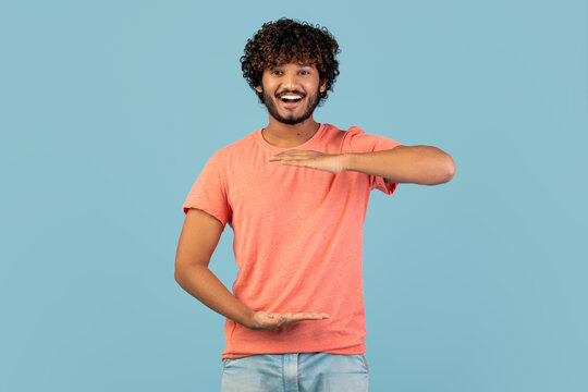 A cheerful young Indian man with curly hair is smiling at the camera while holding something invisible in his hands. He is wearing a pink t-shirt and is set against a blue studio background.