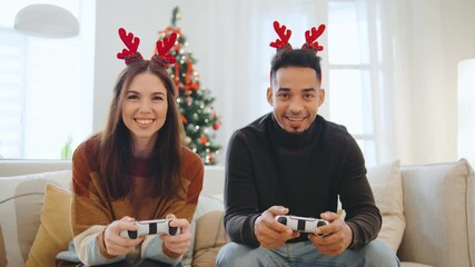 Happy multiracial couple wears reindeer antlers while playing video games together in their living room, celebrating Christmas and New Year with festive decor around them. - Powered by Adobe