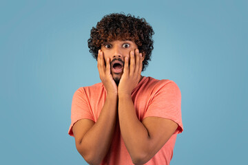 A curly-haired, bearded eastern man is touching his face with both hands. His mouth is open in a...
