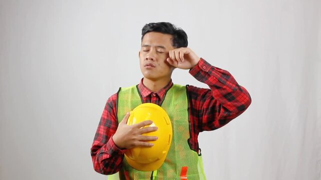 A tired construction worker holding a helmet wipes the sweat from his forehead with his hand.