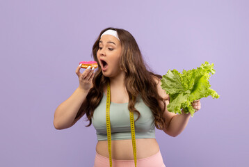 A hungry plus size young female in sportswear enjoys eating a donut while holding a green salad....