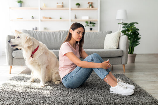 A young woman sits on the floor, looking down in a cozy room. A golden retriever dog is beside her, both appearing calm during the afternoon.