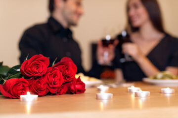 A couple celebrates their love with a candlelit dinner. They toast with drinks while red roses decorate the table, creating a warm, intimate atmosphere.