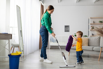 A parent and young child are tidying up their modern living room. The child mimics the parent’s...