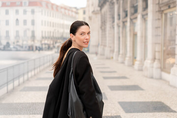Young businesswoman in black coat walking on sidewalk in city. Side view of woman looks back with confident expression. Historic buildings in background create modern urban atmosphere 