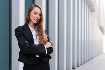 Portrait of smiling woman in business suit standing arms crossed. Young businesswoman with long hair on sidewalk along building in city. She is confidently looking at camera