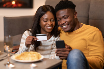 Black couple celebrates Valentine's Day by shopping online using a smartphone and credit card while enjoying a cozy dinner at home. They share smiles and good moments together.
