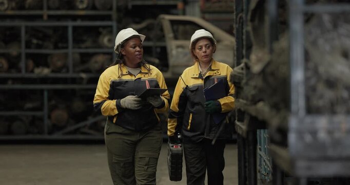 Two female technicians in safety gear inspect, assess used auto parts in factory setting using rugged laptop. Demonstrating technical expertise and teamwork in professional automotive environment
