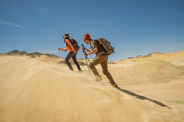 Two travelers with backpacks on their backs, holding trekking poles in their hands, are walking up...