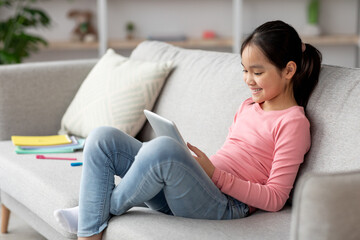 Obraz premium A girl sits comfortably on a gray couch, focused on her tablet. She smiles while wearing a pink long-sleeve shirt and jeans. Colorful school supplies are scattered nearby.