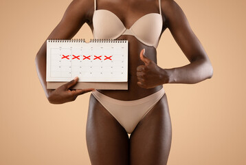 A young African American woman in beige underwear holds a marked calendar and shows a thumb up. She...