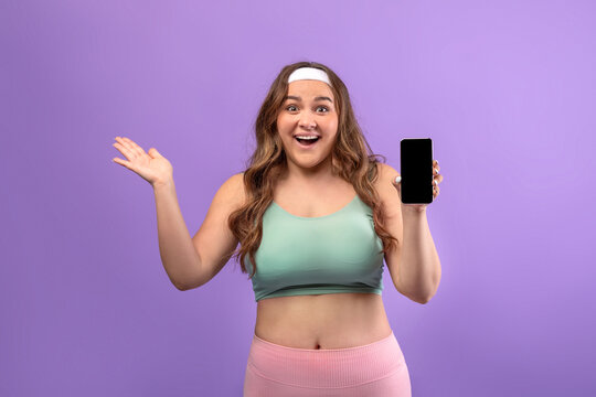 A happy young european plus size woman in sportswear stands against a purple background. She shows a blank phone screen with excitement, promoting a potential amazing offer for a sports app.