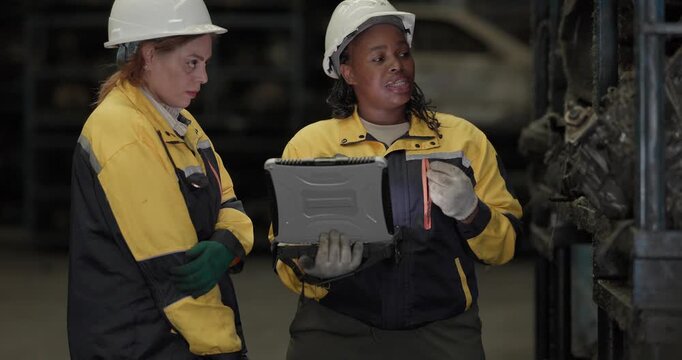 Two female technicians in safety gear inspect, assess used auto parts in factory setting using rugged laptop. Demonstrating technical expertise and teamwork in professional automotive environment
