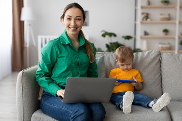 A woman in a green shirt smiles as she works on her laptop. Beside her, a young boy in an orange shirt is focused on his tablet. They are in a bright, modern living room.