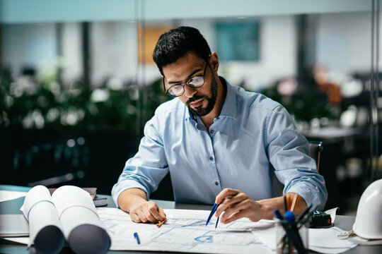 A focused man sketches designs on blueprints at his workspace, surrounded by natural light and plants, showcasing a modern office environment.