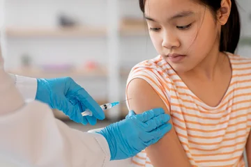 Gardinen Musik A young girl sits calmly while a healthcare worker administers a vaccine to her arm. The clinic is well-lit and organized, focusing on child healthcare.  © Prostock-studio