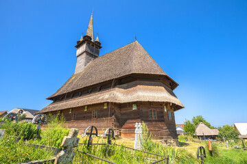 Historic wooden Maramures church of Saint Nicholas in Budesti Josani, Romania, standing under a clear blue sky, surrounded by green grass and traditional cemetery crosses. Maramures of 1600s