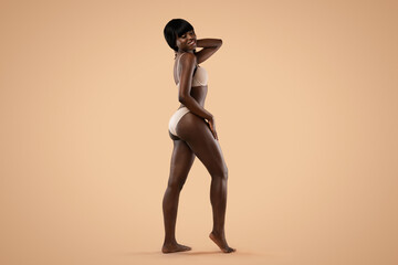 In a colorful studio setting, a confident african american model showcases her shape in beige lingerie. Her striking pose highlights her fit body and long legs in a playful manner.