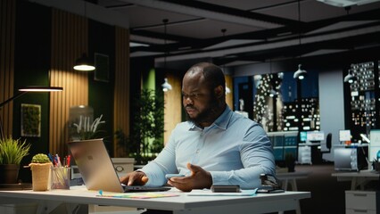 Black businessman sighing and checking messages on mobile phone, feeling drained and frowning after being distracted by a notification in modern office. Workplace challenges at night.
