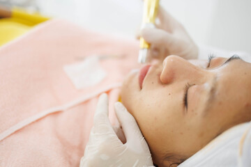 A woman is getting a facial treatment with a gold tipped instrument. The woman is wearing a white hat and a white gown. The woman is smiling and she is relaxed
