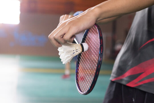 The classic stance for a badminton serve, showing the player's hand holding the white shuttlecock at the moment of release, ready for the red-framed racket to strike on the green court.