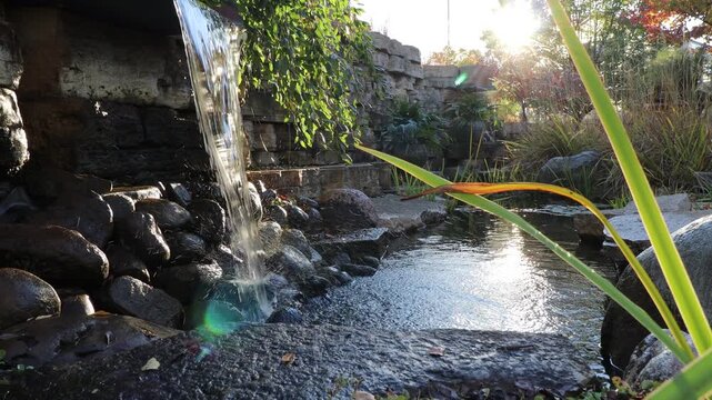 4K tracking video of manmade waterfall in landscaping of a park with the sun high in the sky and green vegetation all around.