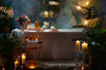 A woman relaxes in a bubble bath, holding a golden bottle amidst plants and candlelight, enjoying aromatherapy and creating a tranquil home spa experience.