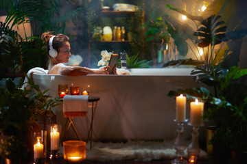 Woman relaxing in a bubble bath wearing headphones and using a tablet, surrounded by plants and candles, embodying self-care and tranquility.
