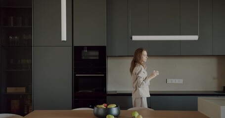 Woman in pajamas walks through her modern kitchen and opens a cupboard. The kitchen is decorated in a minimalist style with dark gray cabinets and a light wood table. She holds a glass of water.