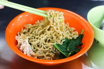 Eating chicken noodles with chopsticks. The dish features wavy yellow noodles topped generously with tender, shredded chicken, fresh green vegetables, and a sprinkle of chopped green onions.