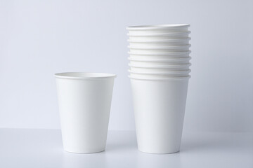 A single white paper cup stands next to a neatly stacked pile of several identical cups on a clean, white background. Suitable for mockups, consumer products, or other concepts.