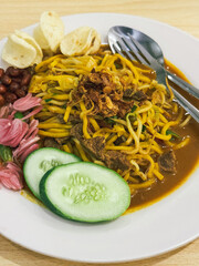 A plate of traditional Indonesian Mie Aceh served with rich spicy curry sauce, beef, cucumber slices, peanuts, crackers, and fried shallots on top.