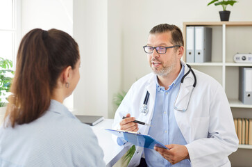 Obraz premium Portrait of a young woman patient in office listening to recommendations of mature male doctor sitting at the desk in clinic during medical examination. Health care and medicine concept.