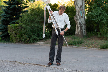  A man in a white shirt and dark trousers carefully unfolds a tripod on a paved area outdoors, preparing it for telescope assembly near stone columns and trees
