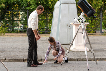  A men in a white shirt demonstrates a telescope mount component to a young student with glasses, who observes carefully during an outdoor astronomy lesson near observatory buildings