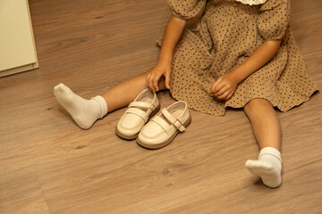 Young girl wearing a brown polka dot dress sits on a wooden floor with white socks, holding a pair of beige shoes beside her, suggesting a moment of dressing or preparation indoors