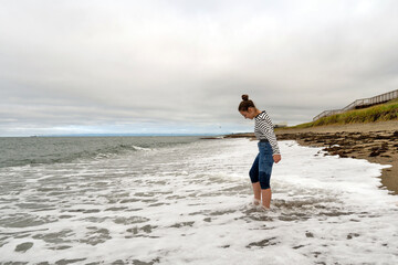 Girl in striped shirt and rolled jeans plays barefoot at the shoreline, stepping into the foamy surf as waves wash around her legs beneath a cloudy coastal sky. Sea of Okhotsk Sakhalin Island