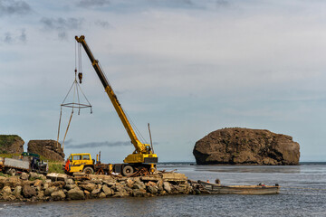 Industrial crane lifting equipment near rocky coastal island. A large yellow crane operates on a rocky shore beside trucks and boats, while a massive rocky island rises in the background, combining in