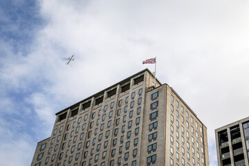 Tall office building with national flag on rooftop and airplane flying in cloudy blue sky