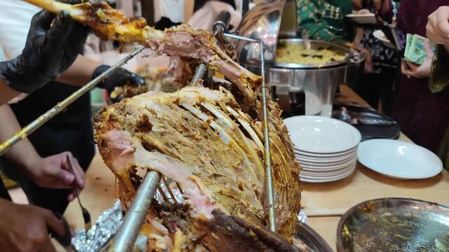 A close-up shot of a chef carving roasted lamb at a buffet station. The lamb, cooked on a rotisserie, shows tender meat and exposed ribs while being sliced with a knife.