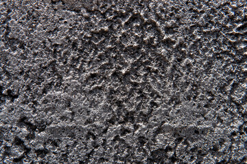 Extreme close-up (macro) of a dark gray or black concrete wall. Bright sunlight creates harsh shadows, highlighting the coarse, gritty, and rough stucco texture. Perfect industrial or grunge backgroun