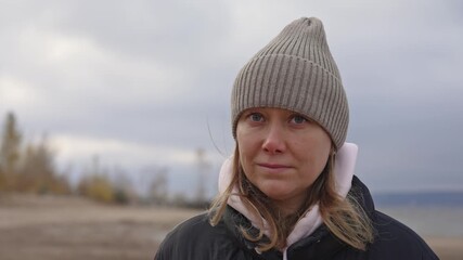 Pensive lady enjoying windy seaside day, contemplative female walking along overcast autumn shoreline, tranquil woman with beanie and earmuffs observing seasonal coastal landscape under gray sky