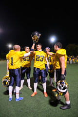 American football players celebrating a win, raising a helmet on a floodlit field at night